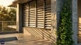 Close-up architectural detail of west-facing first floor balcony showing horizontal aluminium louvre fins, deep chajja projection, rough Kota stone column cladding, and vertical green planter strip