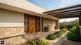 Close-up architectural detail of a south-facing elevation with a deep verandah pergola, horizontal design bands, textured plaster finish, and a lush planted entrance garden