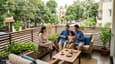 Indian family relaxing on the cool north-facing first-floor balcony lounge of their 30x40 G+3 home, morning light, comfortable seating, green neighbourhood visible in background