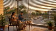Indian couple enjoying evening chai on west-facing first floor balcony with golden hour light filtering through horizontal louvre screen, terracotta plant pots, and soft urban street view below