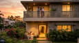 Warm evening scene of a multigenerational Indian family at their west-facing G+1 home — parents relaxing on the ground floor garden, young family on the first floor balcony